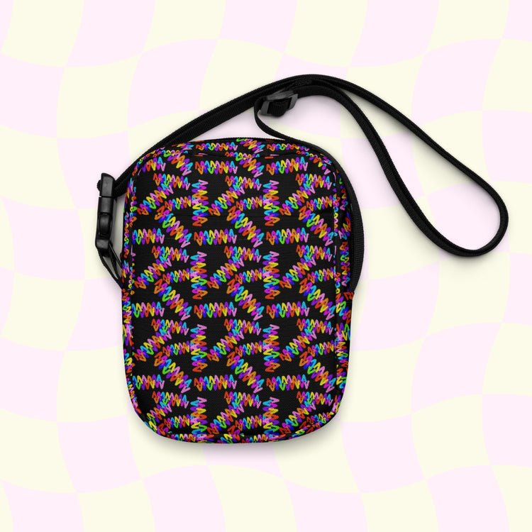 The Electric Zigzag Mini Crossbody Bag by My Favourite Colour is Rainbow features a vibrant electric zigzag print with interlocking red, blue, green, and yellow shapes on a black background. It includes adjustable straps for a perfect fit and has a pale checkered design backdrop.