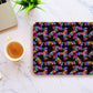 The Electric Zigzag Mouse Pad by My Favourite Colour is Rainbow features a colorful abstract pattern of zigzag lines on black, placed on a marble table next to coffee, a laptop, and a small green plant.