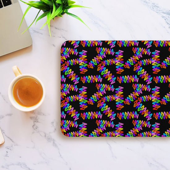 The Electric Zigzag Mouse Pad by My Favourite Colour is Rainbow features a colorful abstract pattern of zigzag lines on black, placed on a marble table next to coffee, a laptop, and a small green plant.