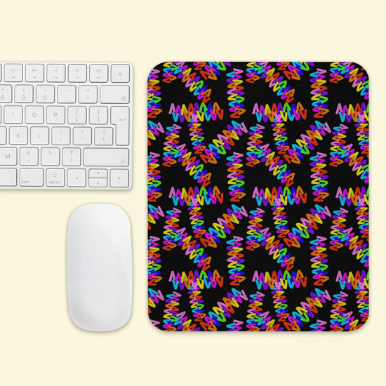 The Electric Zigzag Mouse Pad by My Favourite Colour is Rainbow features vibrant pink, orange, yellow, green, and blue zigzags on a black background, sitting beside a white keyboard and mouse on a light yellow surface.