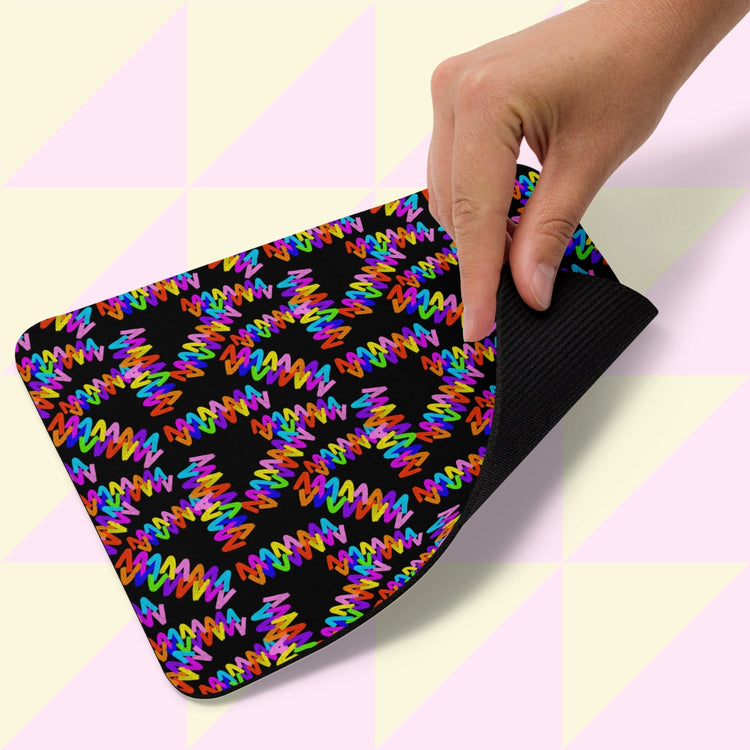 A hand lifts the corner of a My Favourite Colour is Rainbow Electric Zigzag Mouse Pad, revealing a colorful zigzag pattern on a black surface. The background showcases a light pink and cream geometric triangle pattern.