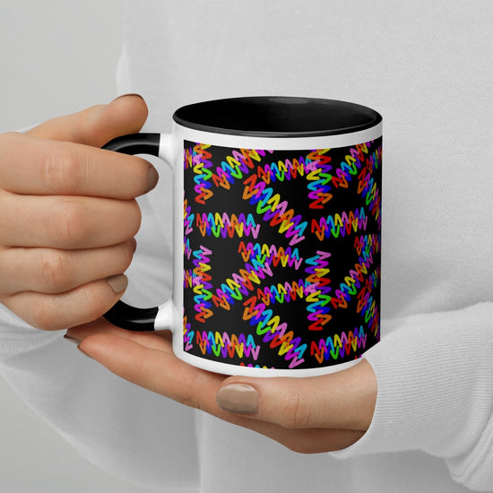 Hands clasp the Electric Zigzag Mug by My Favourite Colour is Rainbow, a ceramic mug boasting a vivid zigzag pattern on a black background. Overlapping multicolored lines deliver an energetic design, ideal for lively coffee breaks. The person wears a white long-sleeve shirt.