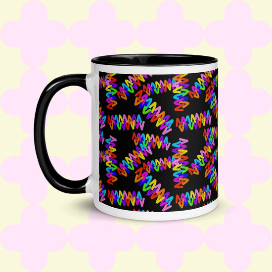 The Electric Zigzag Mug by My Favourite Colour is Rainbow is a ceramic mug with a black handle and rim, featuring a vibrant zigzag pattern in red, blue, green, and yellow over a black background. Its light yellow backdrop displays a subtle pink quatrefoil design—ideal for brightening your coffee break.