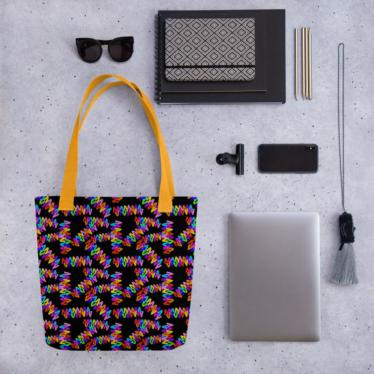 A flat lay on a grey surface: the Electric Zigzag Tote Bag by My Favourite Colour is Rainbow with yellow handles, sunglasses, books, a phone, a laptop, pens, a notebook, a black clip, and a decorative tassel.