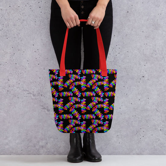 A person carries the Electric Zigzag Tote Bag from My Favourite Colour is Rainbow, featuring spacious, colourful geometric patterns and vivid red handles, as they stand confidently in black pants and boots against a textured grey wall.