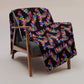 The My Favourite Colour is Rainbows Electric Zigzag Throw Blanket, adorned with a multicolored zigzag pattern on a neutral beige background, covers a wooden chair with a dark gray cushion over its backrest and seat.