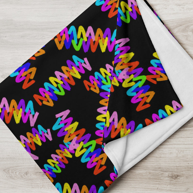 The Electric Zigzag Throw Blanket by My Favourite Colour is Rainbow features a vibrant zigzag pattern in purple, pink, red, orange, yellow, green, and blue on a black background with white reverse. It’s crafted from soft fabric and neatly folded on a light wooden surface.