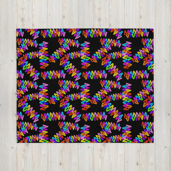 The Electric Zigzag Throw Blanket by My Favourite Colour is Rainbow showcases colorful zigzag patterns on a black background, creating an energetic look with multicolored lines against a soft fabric, beautifully contrasting with a light wooden surface.