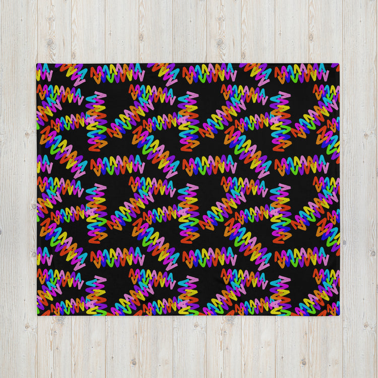 The Electric Zigzag Throw Blanket by My Favourite Colour is Rainbow showcases colorful zigzag patterns on a black background, creating an energetic look with multicolored lines against a soft fabric, beautifully contrasting with a light wooden surface.