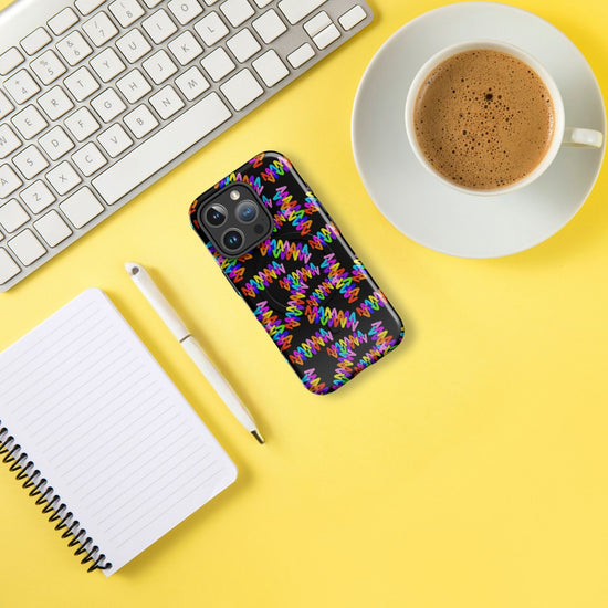 The Electric Zigzag MagSafe iPhone Case by My Favourite Colour is Rainbow, featuring a colorful geometric pattern and shock-absorbing liner, rests next to a white keyboard, coffee cup on a saucer, spiral notebook, and pen—all on a bright yellow surface.