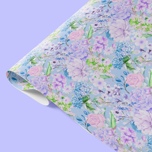 Floral Wrapping Paper Sheets by My Favourite Colour is Rainbow feature pastel flowers like pink roses, purple hydrangeas, and green leaves on a light blue background. Partially folded, the sheet of wrapping paper is displayed on a lavender surface.