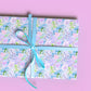 A gift wrapped in My Favourite Colour is Rainbows Floral Wrapping Paper Sheets, showcasing blue, pink, and green flowers with a light blue ribbon on a solid pink surface.