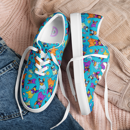 A pair of Frenchie Fetch, Set, Match Canvas Sneakers from My Favourite Colour is Rainbow, showcasing cartoon cats and yarn on a blue background with white soles and laces, sit atop a peach knit sweater and blue jeans.