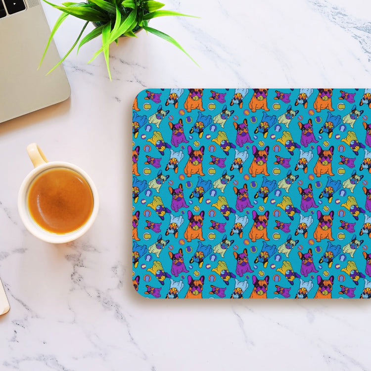 A marble desk holds the Frenchie Fetch, Set, Match Mouse Pad by My Favourite Colour is Rainbow, showcasing cartoon dogs in playful poses on a bright blue background. Nearby sit a laptop, a green plant, and a cup of coffee.