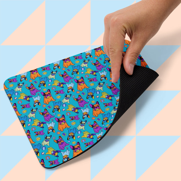 A hand lifts the corner of the My Favourite Colour is Rainbows Frenchie Fetch, Set, Match Mouse Pad, showcasing its playful pattern of cartoon dogs and balls on a blue background. The underside reveals a light blue and peach geometric design.