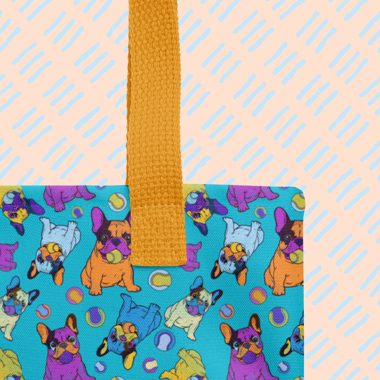 A closeup of the Frenchie Fetch, Set, Match Tote Bag by My Favourite Colour is Rainbow featuring cartoon French Bulldogs and tennis balls in purple, orange, and blue on a bright blue background.   It has a sturdy yellow strap.