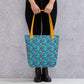 An individual carries the spacious Frenchie Fetch, Set, Match Tote Bag by My Favourite Colour is Rainbow, featuring colourful french bulldog illustrations and yellow handles. They stand confidently in black pants and boots against a textured grey background.