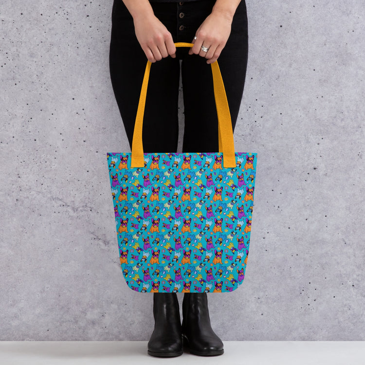 An individual carries the spacious Frenchie Fetch, Set, Match Tote Bag by My Favourite Colour is Rainbow, featuring colourful french bulldog illustrations and yellow handles. They stand confidently in black pants and boots against a textured grey background.