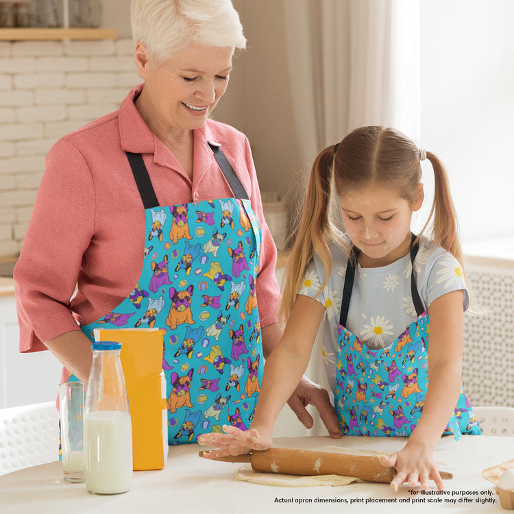 In a joyful kitchen scene, a woman and a young girl are baking together, both wearing My Favourite Colour is Rainbows Frenchie Fetch, Set, Match Apron featuring colourful cartoon french bulldogs. The girl rolls dough as the woman smiles nearby. A box of flour and bottles of milk sit on the table.