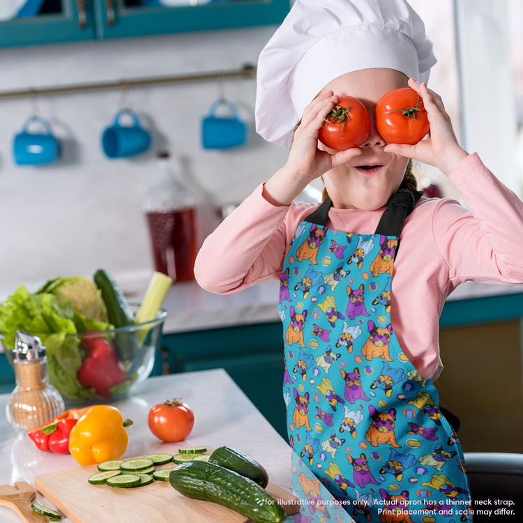 A child playfully holds two tomatoes over their eyes, wearing a vibrant Frenchie Fetch, Set, Match Apron and chefs hat from My Favourite Colour is Rainbow. Cucumbers, a yellow bell pepper, lettuce, and salad dressing grace the counter in this delightful kitchen scene.