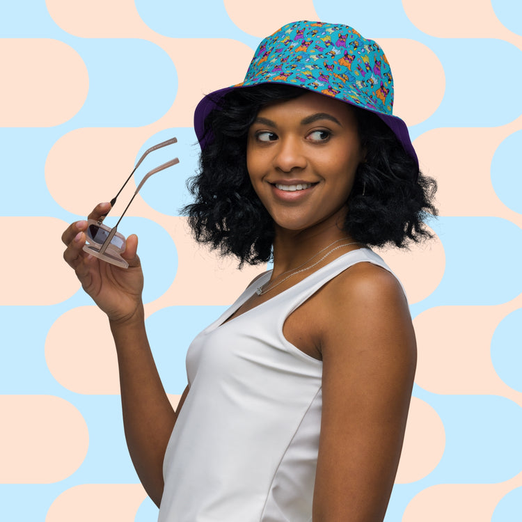 A woman in a white sleeveless top wears the Frenchie Fetch, Set, Match reversible bucket hat by My Favourite Colour is Rainbow. Holding clear glasses and smiling leftward against a pastel geometric background of blue and peach ovals, she showcases this trendy streetwear accessory.