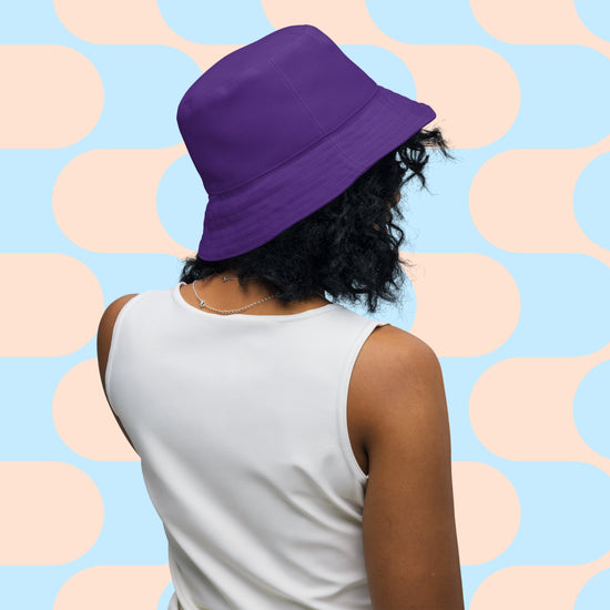 A person with curly hair wears the Frenchie Fetch, Set, Match Reversible Bucket Hat by My Favourite Colour is Rainbow and a sleeveless white top. They stand facing away against a retro geometric pattern in pastel blue and peach tones.