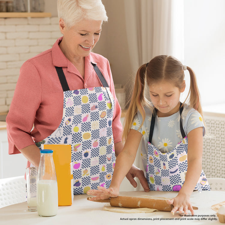 A woman and a young girl, both in vibrant Fruit Salad Aprons by My Favourite Colour is Rainbow, are baking together. The woman smiles as the girl rolls out dough on a table filled with baking ingredients like milk and flour.