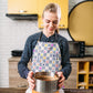 Wearing her Fruit Salad Apron by My Favourite Colour is Rainbow, a woman holds a springform pan with a cake in a modern kitchen featuring a sleek oven and vibrant yellow cabinet, creating a cheerful ambiance.