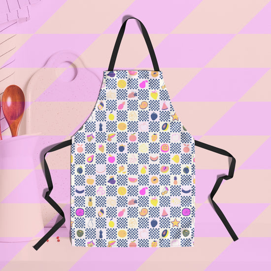 The Fruit Salad Apron by My Favourite Colour is Rainbow showcases a vibrant geometric design with colourful fruit symbols. Made from premium fabric, it has black ties.  The background features wooden kitchen utensils and a chopping board on the left.