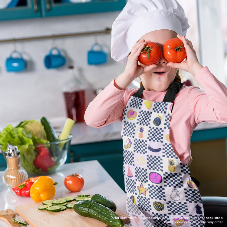 A child in a chefs hat and My Favourite Colour is Rainbows Fruit Salad Apron holds two tomatoes over their eyes like glasses. In the bright kitchen, with its playful atmosphere, cucumbers and peppers rest on a cutting board, showcasing delightful kitchen attire.