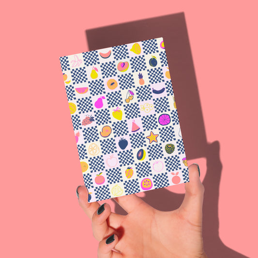 A hand holds the Fruit Salad Greeting Card from My Store, showcasing a checkerboard design with vibrant papaya and pineapple illustrations among geometric shapes. The pink background casts a shadow on the card, and black-painted nails are visible.