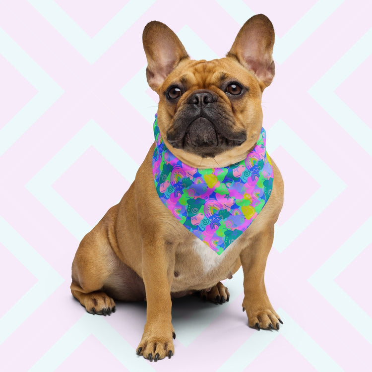 A brown French Bulldog sits in front of a pink and white geometric background, wearing the vibrant Funfetti Bandana by My Favourite Colour is Rainbow. The bandana features an abstract pattern with vibrant swirls, rainbows and stars in blue, pink, purple and green.