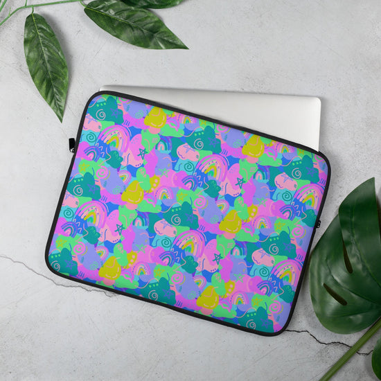 The 15" Funfetti Laptop Sleeve by My Favourite Colour is Rainbow, with a vibrant design of pink, green, blue, and purple rainbows, stars, swirls and abstract shapes, has a silver laptop coming out of it.  It is displayed on a light gray surface, bordered by large green leaves.  The print is on only one side of the laptop sleeve, the other side is black.