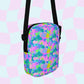 The Funfetti Mini Crossbody Bag by My Favourite Colour is Rainbow features a vibrant psychedelic pattern in pink, blue, green, and purple with a black strap. It beautifully complements the wavy pastel pink and blue background.