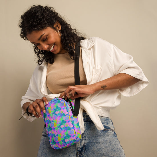 A woman smiles as she reaches into her Funfetti Mini Crossbody Bag by My Favourite Colour is Rainbow, savoring its hands-free ease. Clad in a beige top, white shirt, and blue jeans against a plain backdrop, her outfit complements the bags lively purple and green pattern.