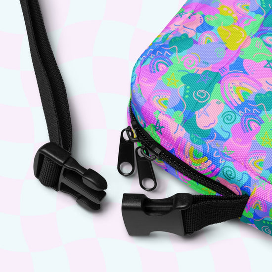 Close-up of the My Favourite Colour is Rainbows Funfetti Mini Crossbody Bag showcasing its abstract pink, green, and blue design. It includes a black zipper with dual sliders and a black strap with a plastic buckle, all on a light checkered background for hands-free convenience.