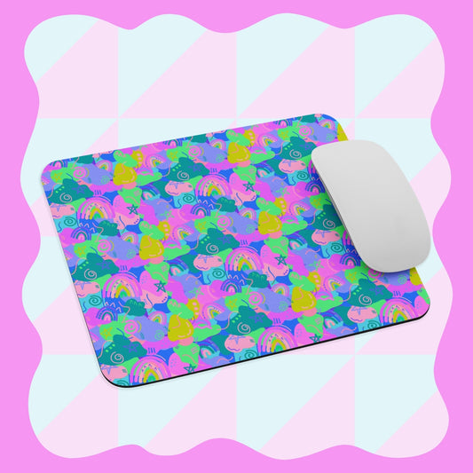 A white wireless mouse sits on the Funfetti Mouse Pad by My Favourite Colour is Rainbow, featuring vibrant unicorns, rainbows, and stars in neon colors against a soft pink and light blue checkerboard background with a wavy border.