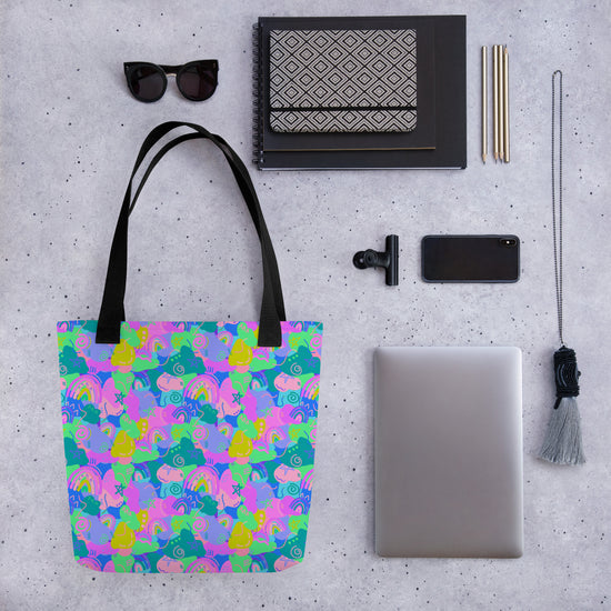 Flat lay of the Funfetti Tote Bag by My Favourite Colour is Rainbow, featuring a colourful pattern of rainbows, clouds, and abstract shapes in pink, green, and blue hues, with sunglasses, a patterned notebook, pens, a smartphone, a laptop, a tassel accessory, and a clip-on camera lens.