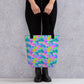 Person holding a Funfetti Tote Bag by My Favourite Colour is Rainbow, showcasing a colourful pattern of rainbows, clouds, and abstract shapes in pink, green, and blue hues. They wear black pants and ankle boots against a light speckled background.