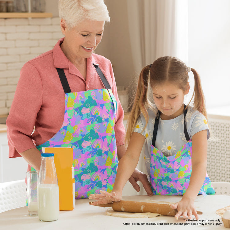 A woman and a young girl, both wearing vibrant Funfetti Aprons by My Favourite Colour is Rainbow, smile as they bake together. The woman watches as the girl rolls dough with a rolling pin on the table surrounded by milk and flour.  The aprons are brightly coloured in shades of green, purple, pink and blue and features rainbows and abstract shapes.