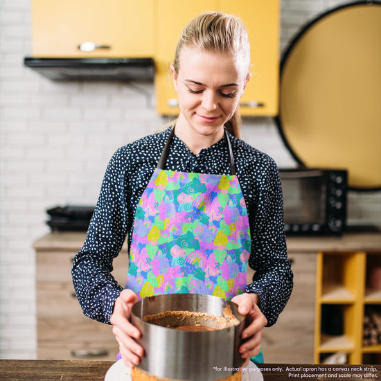 A person wearing a Funfetti Apron by My Favourite Colour is Rainbow prepares dessert in the kitchen. They hold a cake mold, standing before a counter with cabinets and a microwave in the background.  The apron is brightly coloured in shades of green, purple, pink and blue and features rainbows and abstract shapes.