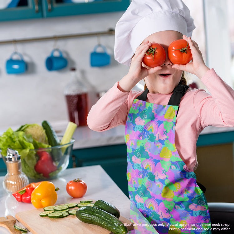 A child in a Funfetti Apron by My Favourite Colour is Rainbow playfully holds two tomatoes over their eyes during a lively cooking adventure, with fresh cucumbers and bell peppers nearby.  The apron is brightly coloured in shades of green, purple, pink and blue and features rainbows and abstract shapes.