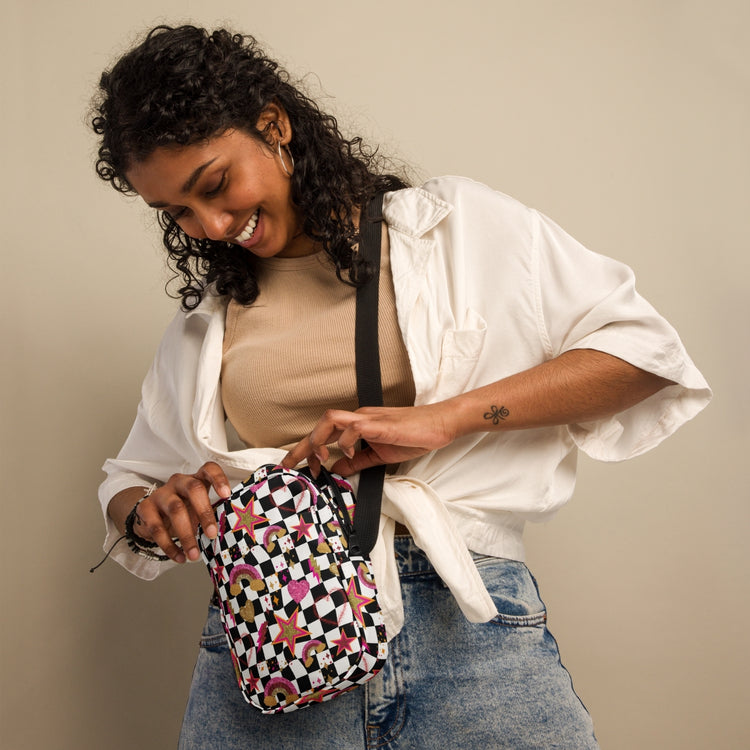 A smiling woman with curly hair, dressed in a white shirt and jeans, opens the Galaxy Mini Crossbody Bag by My Favourite Colour is Rainbow, featuring a colorful geometric pattern and adjustable straps. The background is neutral.