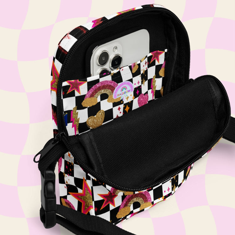 The Galaxy Mini Crossbody Bag by My Favourite Colour is Rainbow features a vibrant, checkered design with stars, rainbows, and hearts. A smartphone is shown in the internal pocket of the partially unzipped bag. 