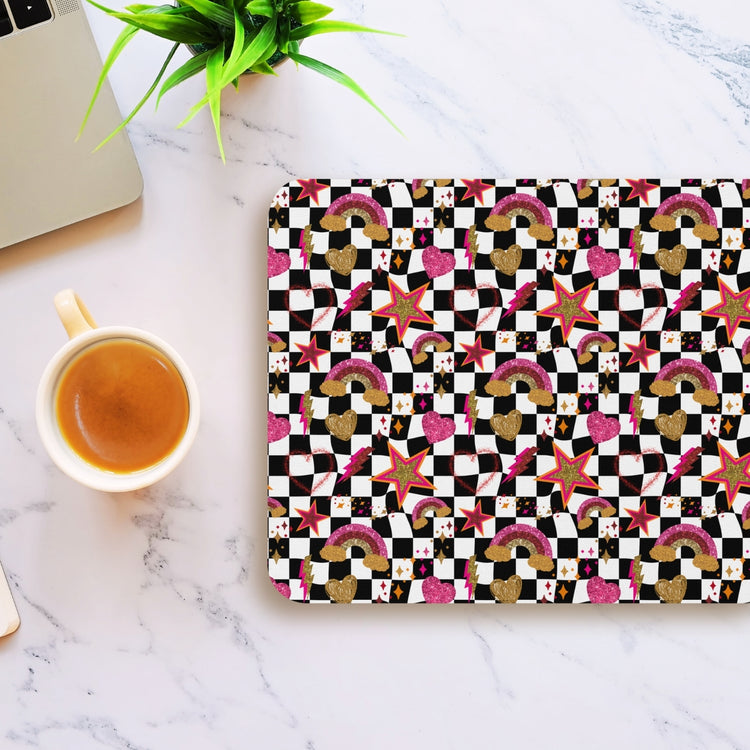 A coffee cup rests on a marble surface next to a Galaxy Mouse Pad by My Favourite Colour is Rainbow, featuring rainbows, stars, hearts, and checkerboard squares. A green plant peeks from above.