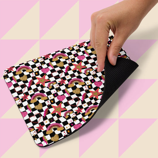 A hand lifts a Galaxy Mouse Pad by My Favourite Colour is Rainbow against a geometric backdrop. The pad showcases playful cupcakes, crowns, stars, rainbows, and checkerboard patterns in black, white, pink, and gold.