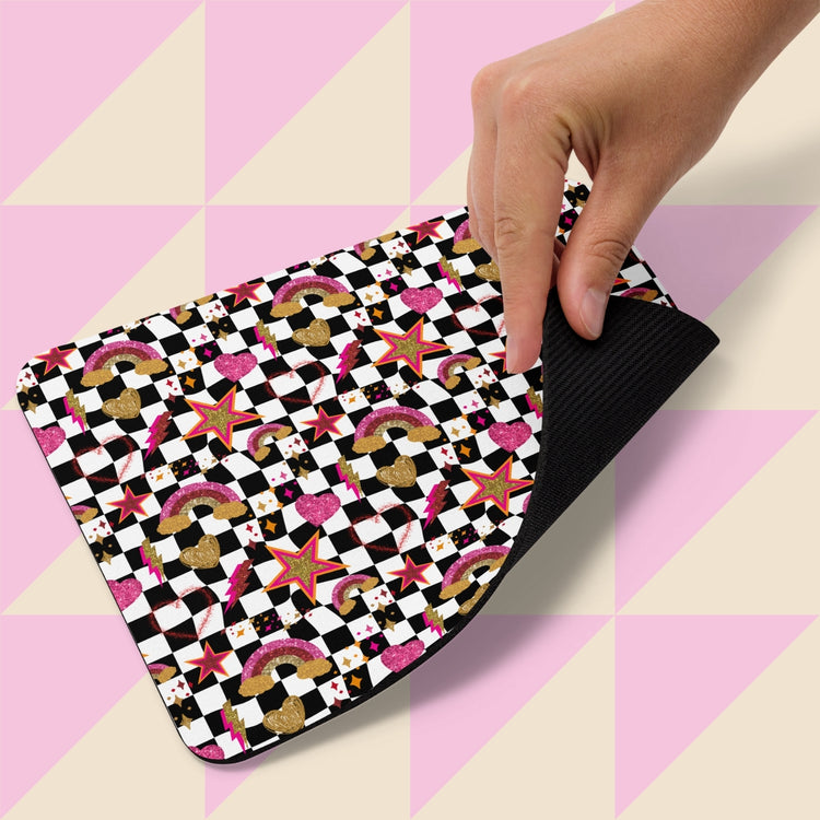 A hand lifts a Galaxy Mouse Pad by My Favourite Colour is Rainbow against a geometric backdrop. The pad showcases playful cupcakes, crowns, stars, rainbows, and checkerboard patterns in black, white, pink, and gold.