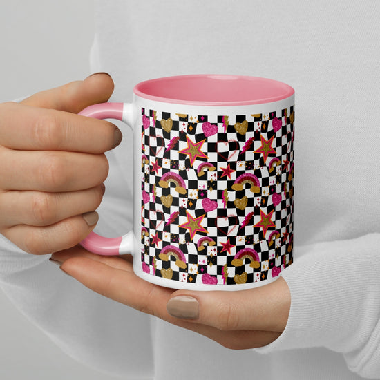 A person with neutral-toned nails holds the My Favourite Colour is Rainbows Galaxy Mug, a white ceramic piece with a pink interior adorned with colorful geometric patterns, rainbows, stars, hearts, and checkerboard designs.