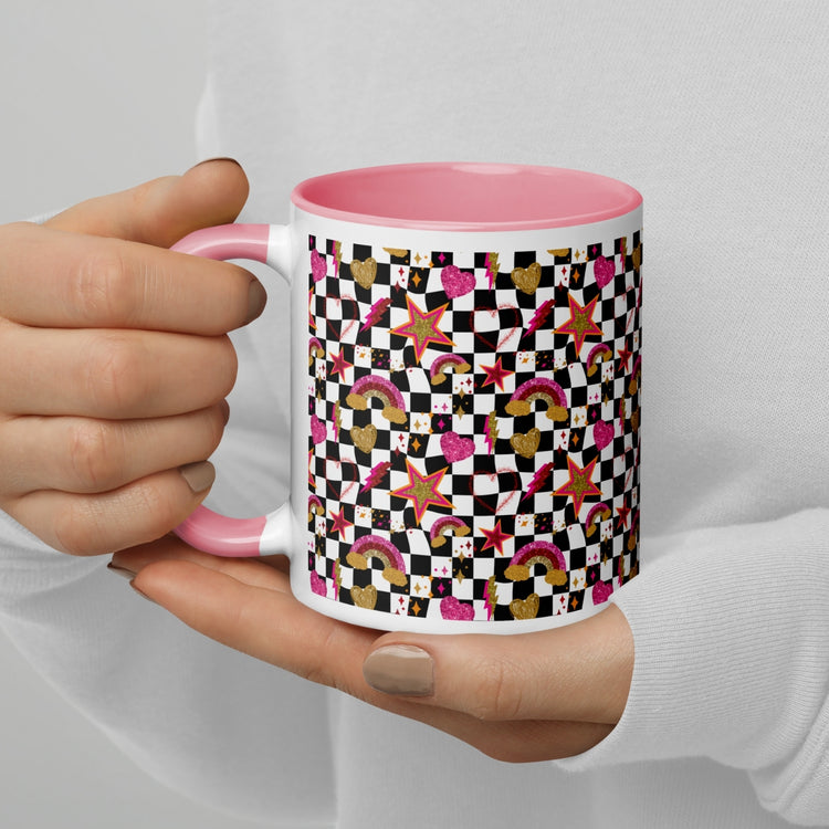 A person with neutral-toned nails holds the My Favourite Colour is Rainbows Galaxy Mug, a white ceramic piece with a pink interior adorned with colorful geometric patterns, rainbows, stars, hearts, and checkerboard designs.