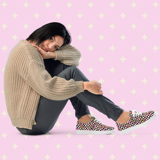 A woman relaxes in a beige knit sweater, dark pants, and My Favourite Colour is Rainbows Galaxy Canvas Sneakers. She rests her head on her knee.  Shown against a soft pink backdrop with white stars.